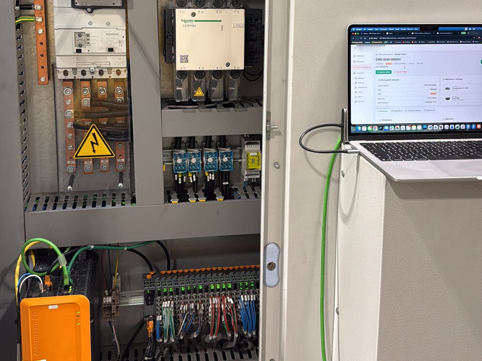 Quadro elettrico aperto con PLC e moduli I/O, accanto un portatile con il software MES su un ordine di produzione